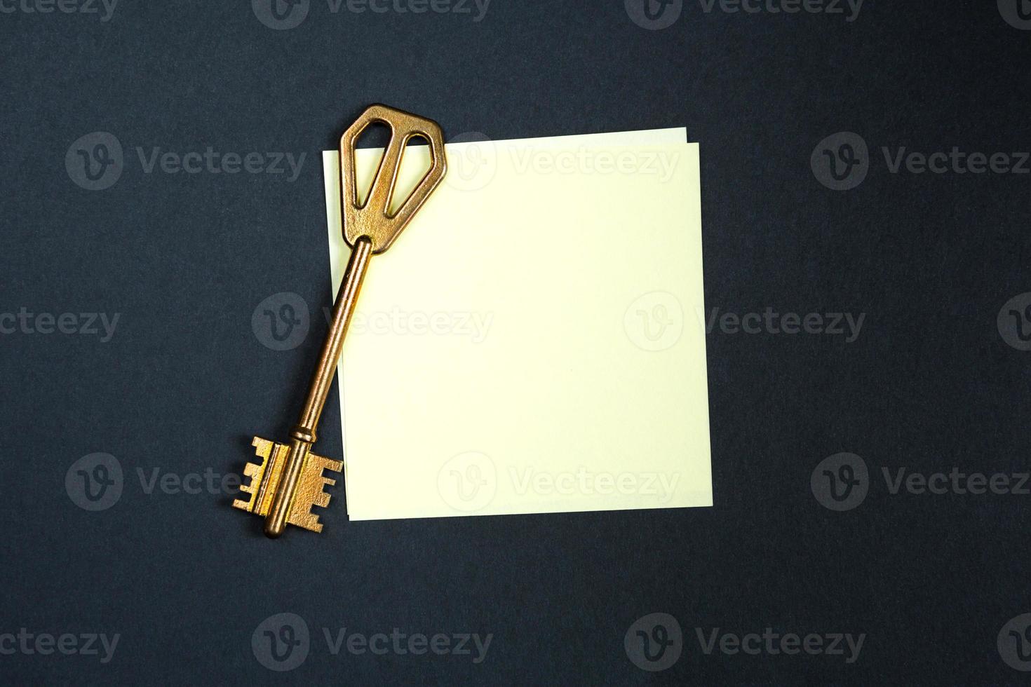 A golden key on a piece of note paper. Wish list, problem solution, idea, decoding, business concept on a black background with a square sheet. Project, rent, booking, household chores. Copy space photo