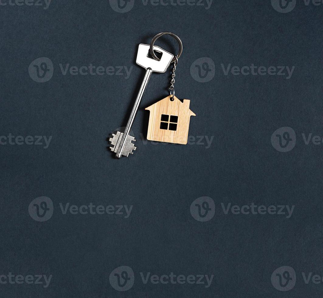Key chain in the shape of wooden house with key on a black background. Building, design, project, moving to new home, mortgage, rent and purchase real estate. Copy space photo