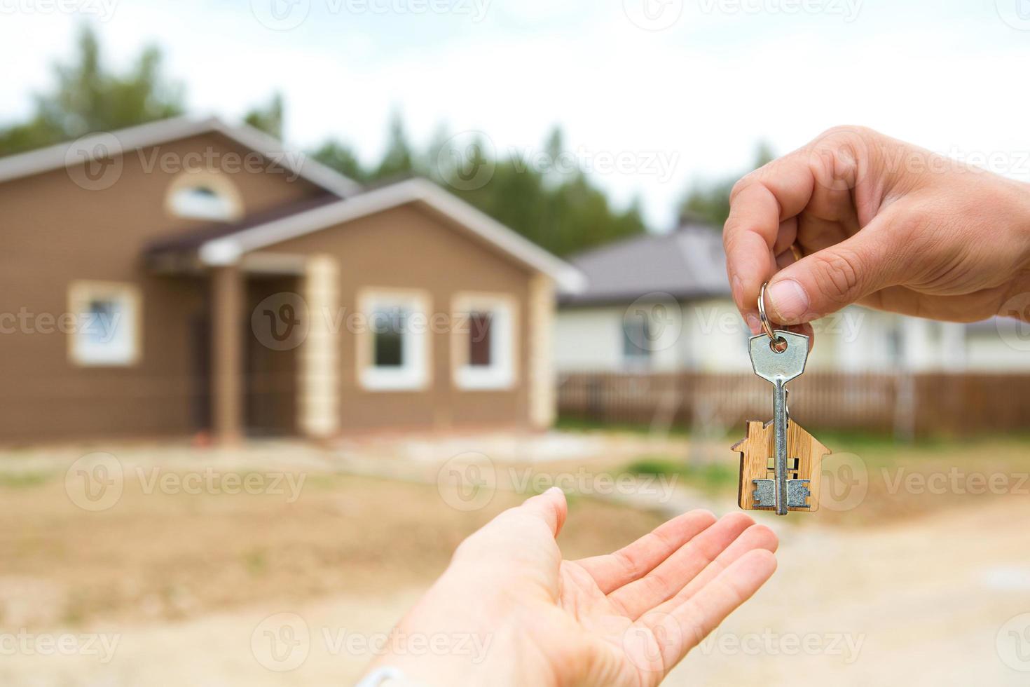 Hand with a key and a wooden key ring-house. Background of fence and cottage. Building, project, moving to a new home, mortgage, rent and purchase real estate. To open the door. Copy space photo