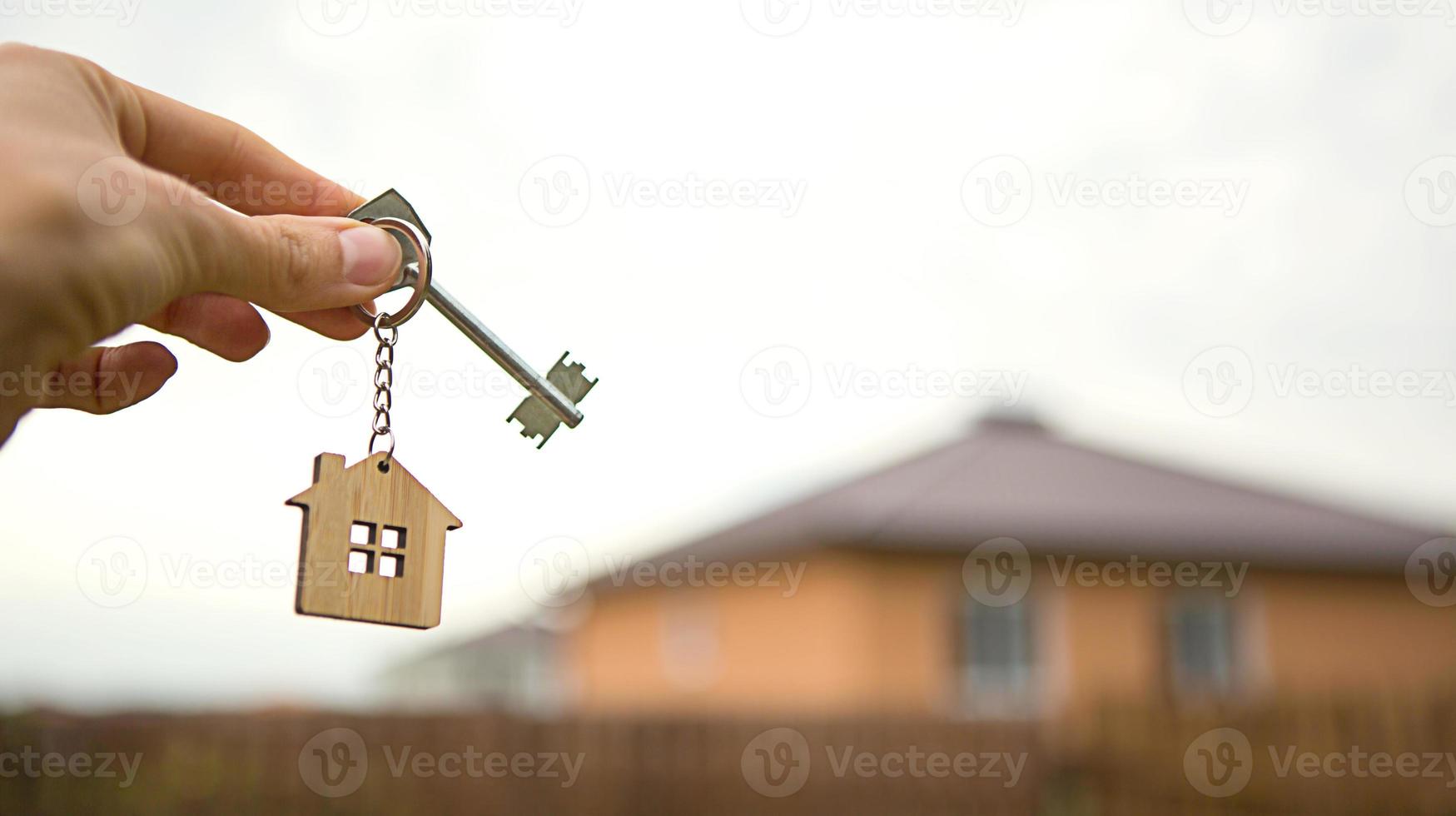 Hand with a key and a wooden key ring-house. Background of fence and cottage. Building, project, moving to a new home, mortgage, rent and purchase real estate. To open the door. Copy space photo