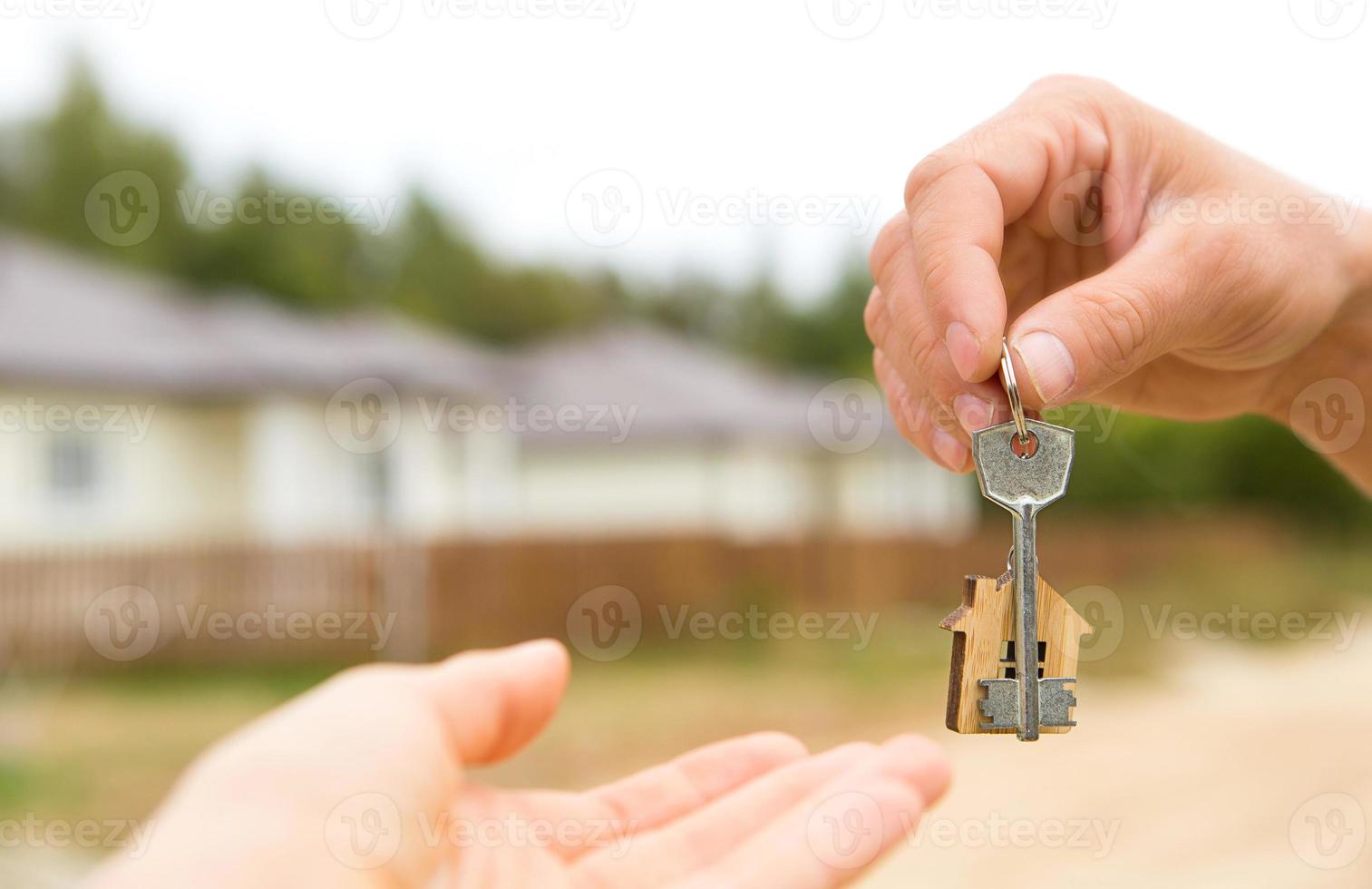 Hand with a key and a wooden key ring-house. Background of fence and cottage. Building, project, moving to a new home, mortgage, rent and purchase real estate. To open the door. Copy space photo
