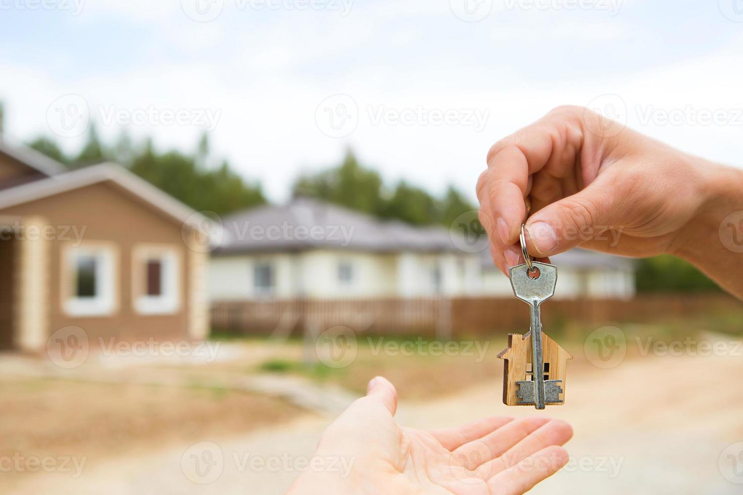 Hand with a key and a wooden key ring-house. Background of fence and cottage. Building, project, moving to a new home, mortgage, rent and purchase real estate. To open the door. Copy space photo