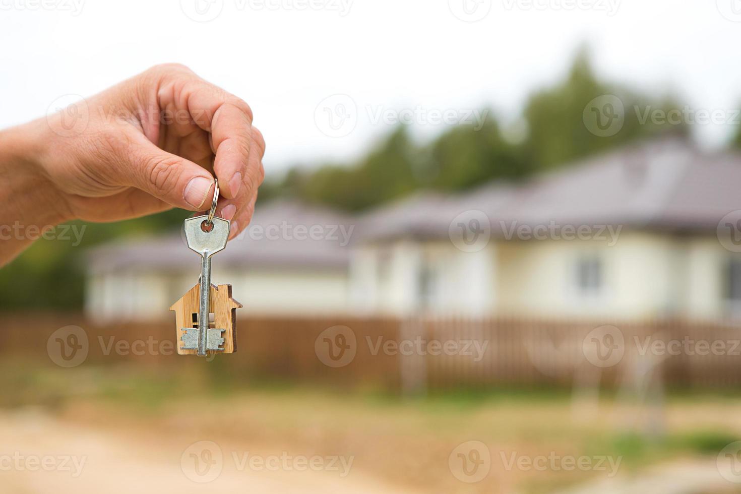 Hand with a key and a wooden key ring-house. Background of fence and cottage. Building, project, moving to a new home, mortgage, rent and purchase real estate. To open the door. Copy space photo