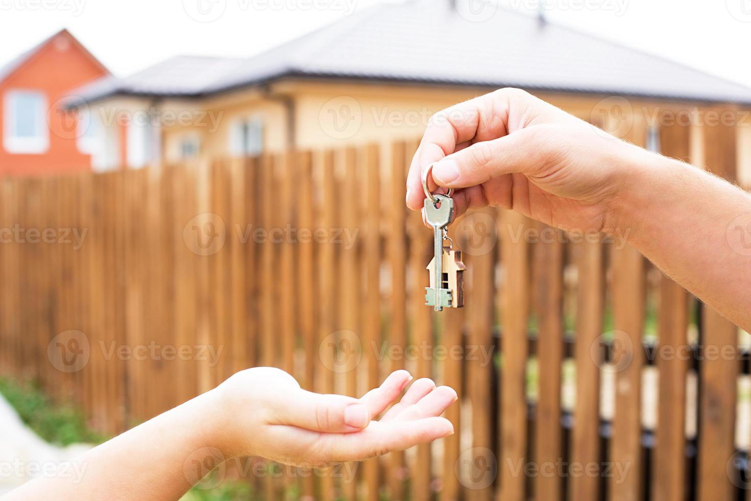 Hand with a key and a wooden key ring-house. Background of fence and cottage. Building, project, moving to a new home, mortgage, rent and purchase real estate. To open the door. Copy space photo