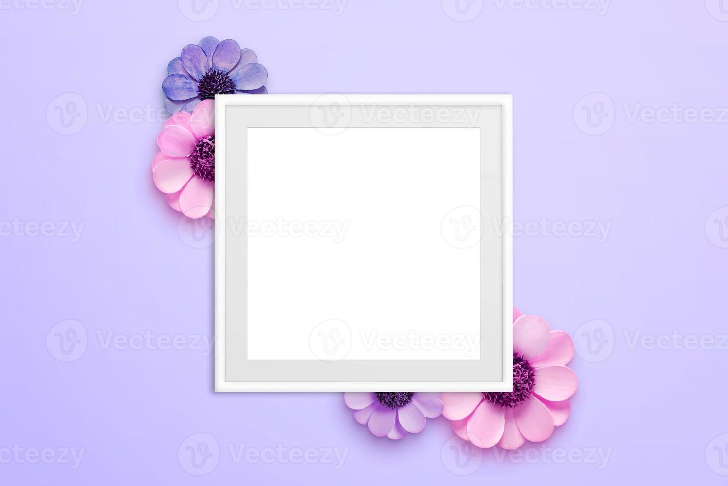 Blank picture frame surrounded with flowers on purple surface. Top view, flat lay composition photo