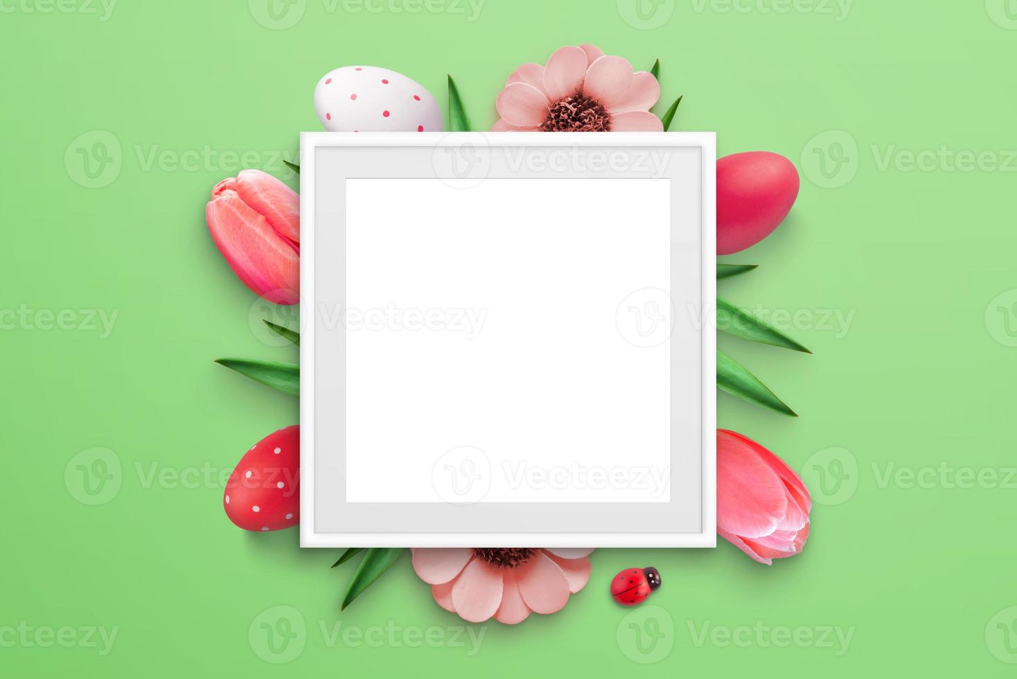 Blank picture frame surrounded by Easter eggs and flowers on green surface. Top view, flat lay composition photo