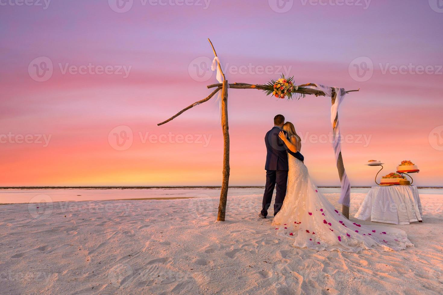The bride and groom under wedding arch on beach. Romantic wedding background. Amazing beach sunset, infinity sea view, romantic clouds sky. Idyllic destination honeymoon photo