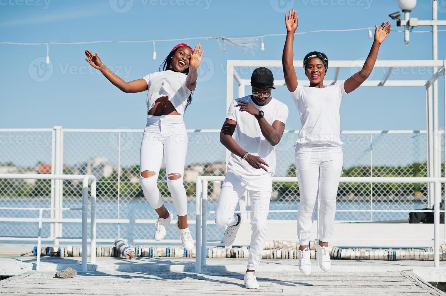 tres elegantes amigos afroamericanos, vestidos con ropa blanca en el muelle de la playa. moda callejera de jóvenes negros. hombre negro con dos niñas africanas. foto