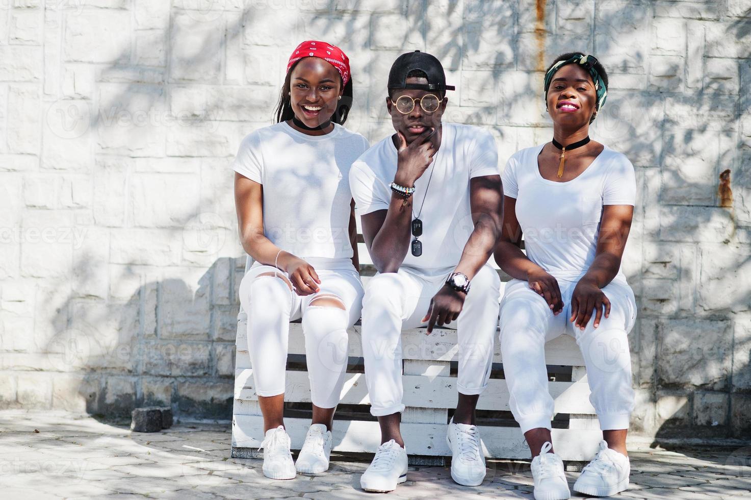 Three stylish african american friends, wear on white clothes sitting on bench. Street fashion of young black people. Black man with two african girls. photo