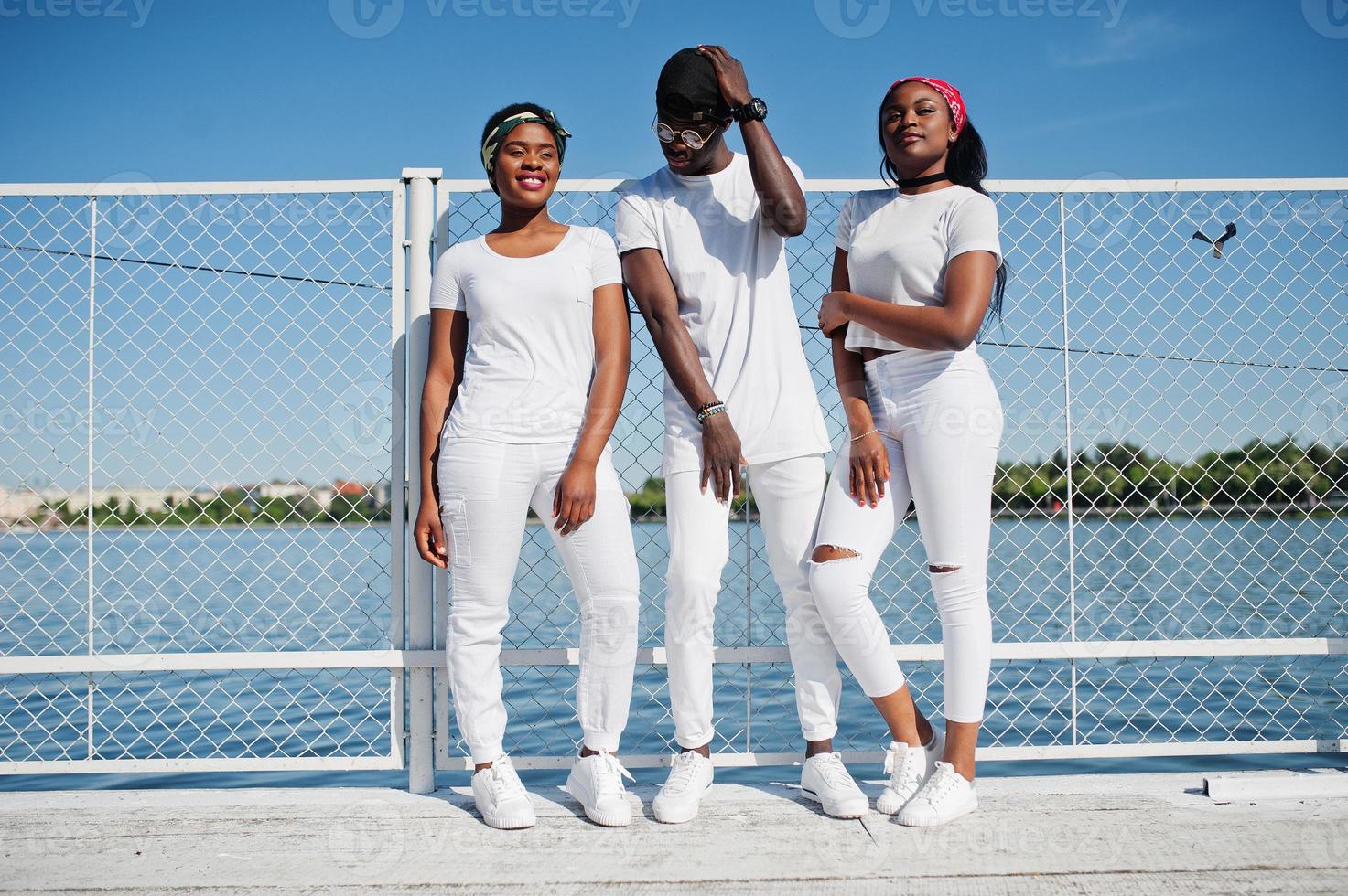 Three stylish african american friends, wear on white clothes at pier on beach against cage. Street fashion of young black people. Black man with two african girls. photo