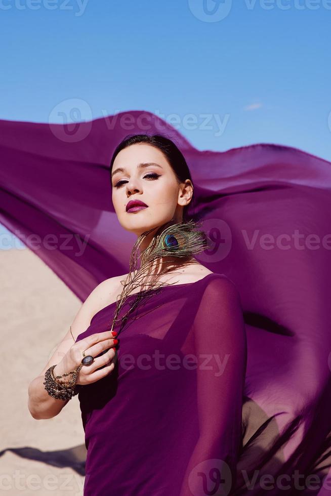 Amazing beautiful brunette woman with the Peacock feather in purple fabric in the desert. Oriental, Indian, fashion, style concept photo