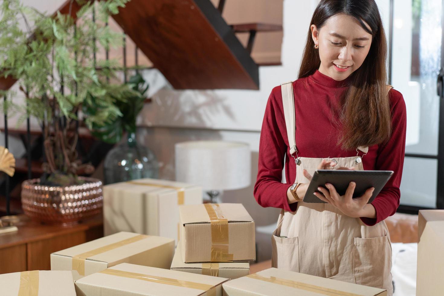 SME small business Asian female entrepreneur or shop owner checking customer order on digital tablet while packing boxes at home. using social for marketing online shopping, e-commerce concept photo