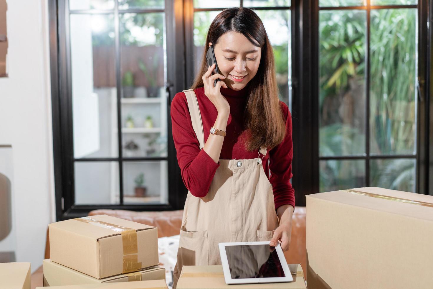 Young Asian shop owner woman calling to customer and checking stock on digital tablet at home. SME small business entrepreneur prepare for shipping product, online store e-commerce or service support photo