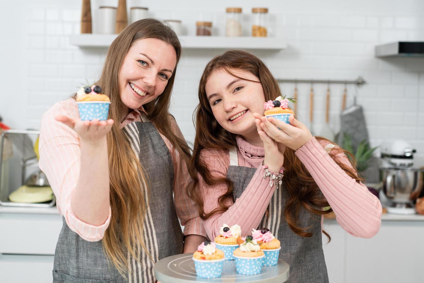 feliz madre e hija mostrando sonrisas y pastelitos caseros mirando la cámara en la cocina. actividad familiar hornear panadería en casa el fin de semana foto
