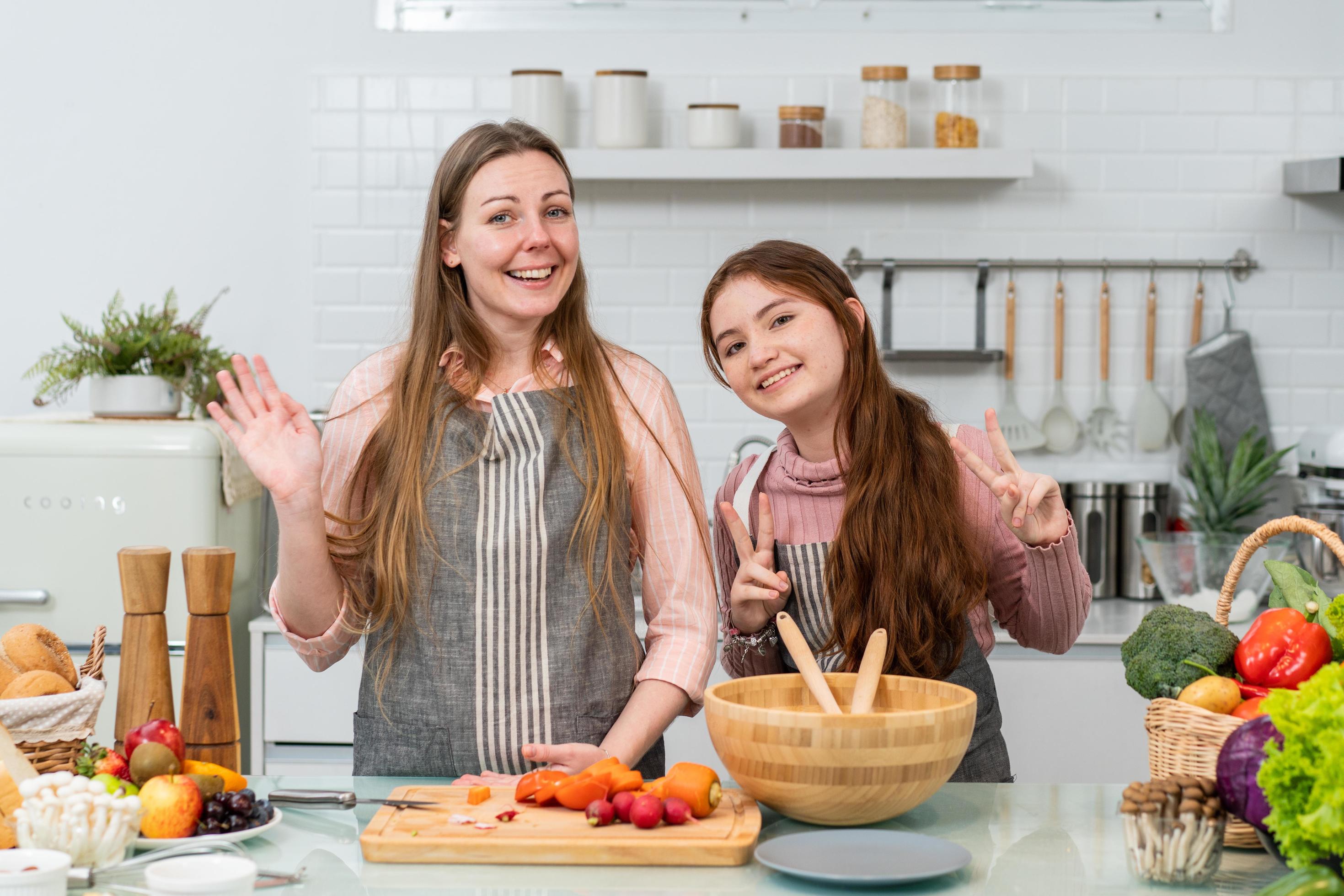 POV of Caucasian mother and daughter greeting to audience showing cooking homemade salad streaming online in the kitchen