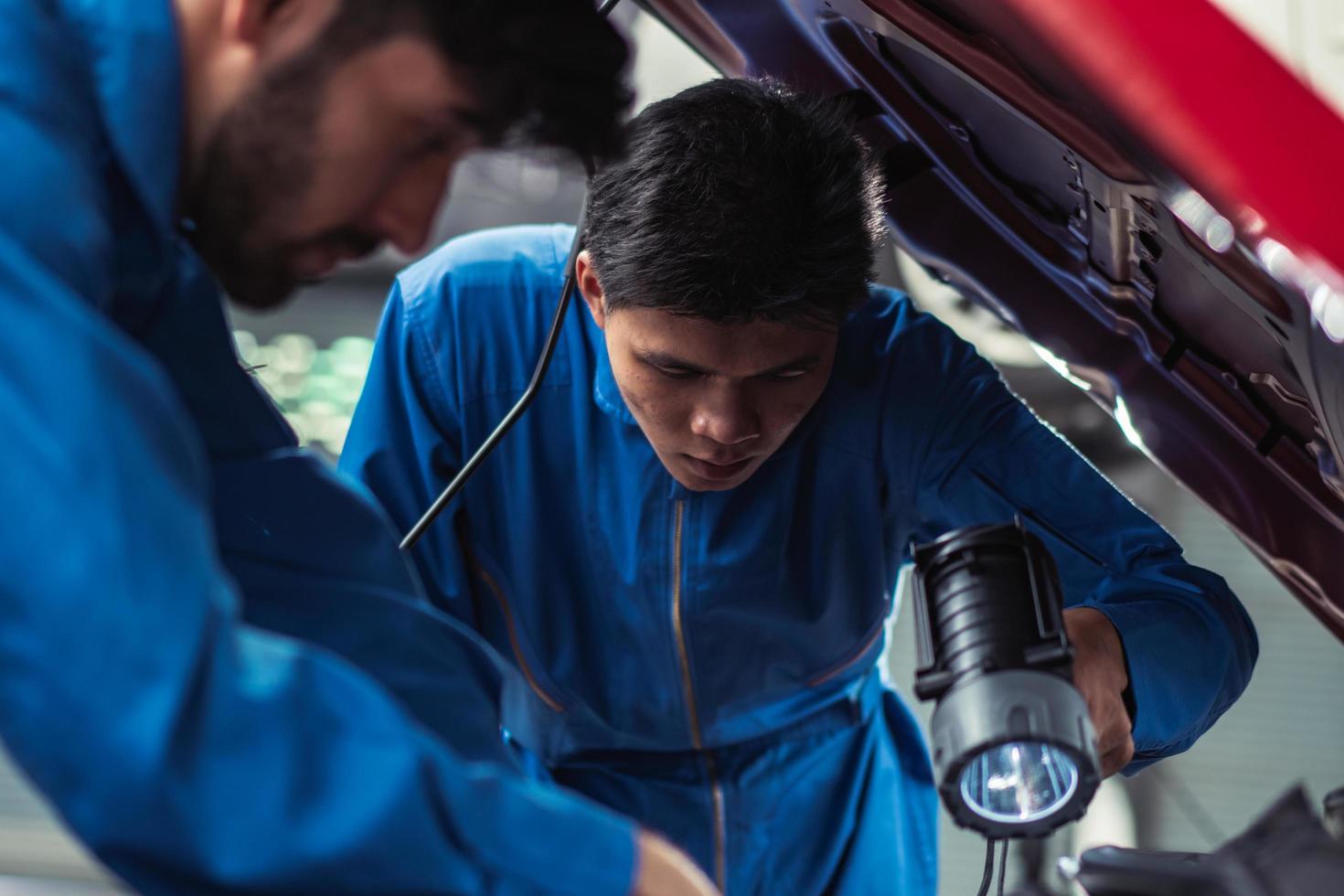 Mechanic shining flashlight to help colleagues repairing car engine at auto garage shop. technician or engineer team checking car damage. after service concept photo