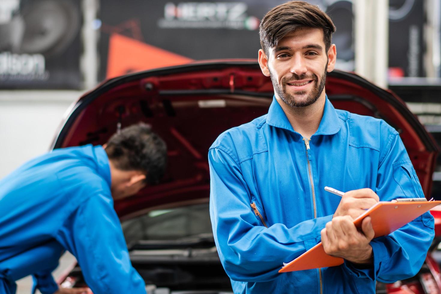 Portrait of Caucasian automotive mechanic man writing on maintenance document clipboard and checking auto damage in auto garage, assistant repairing car, transport business and after service concept photo