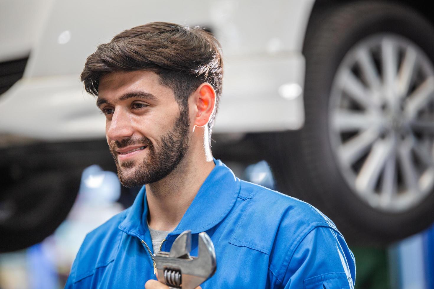 Smiling Caucasian male mechanic holding wrench at auto garage. repair and maintenance career. after service, checking car damage broken part photo