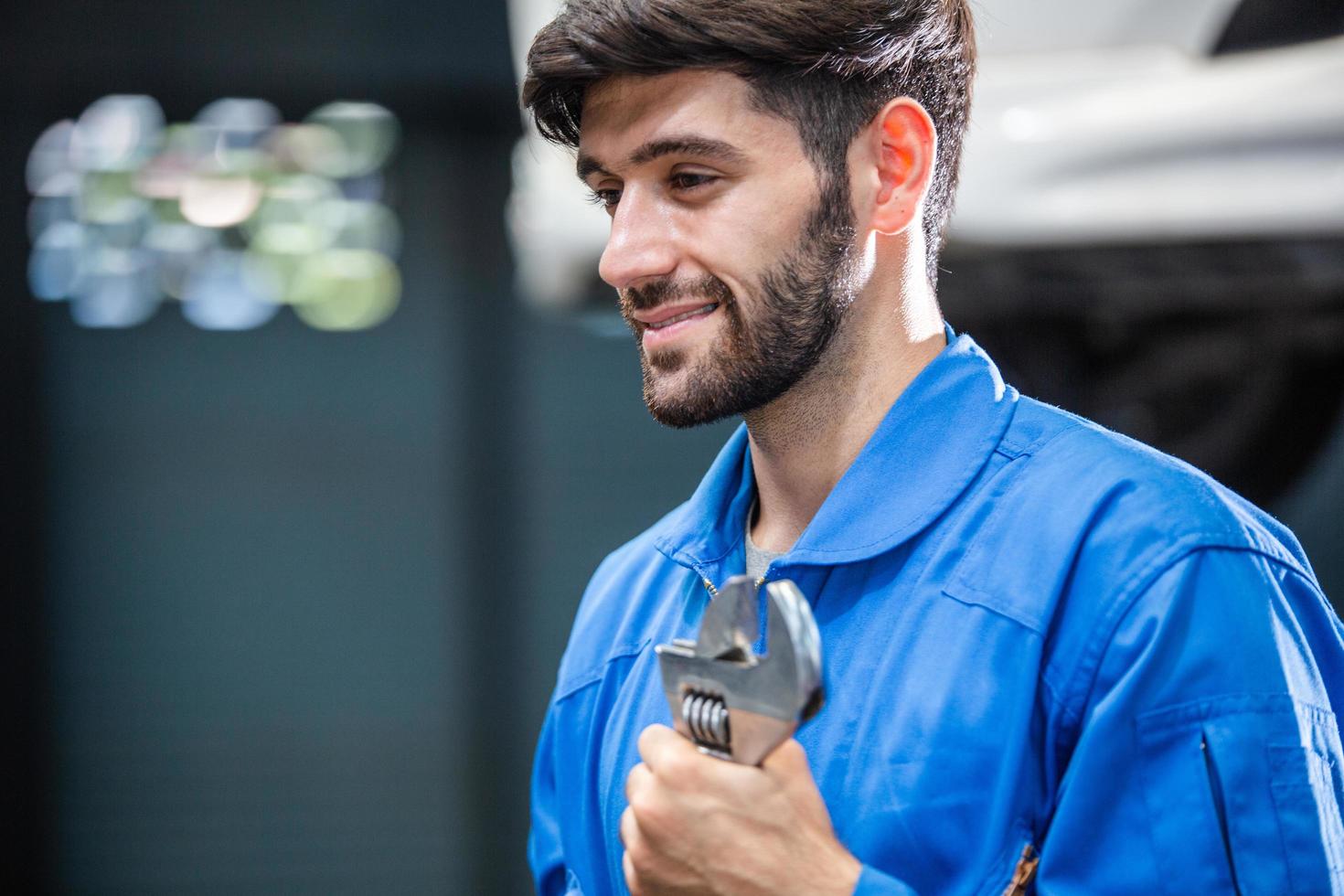 Smiling Caucasian male mechanic holding wrench at auto garage. repair and maintenance career. after service, checking car damage broken part photo