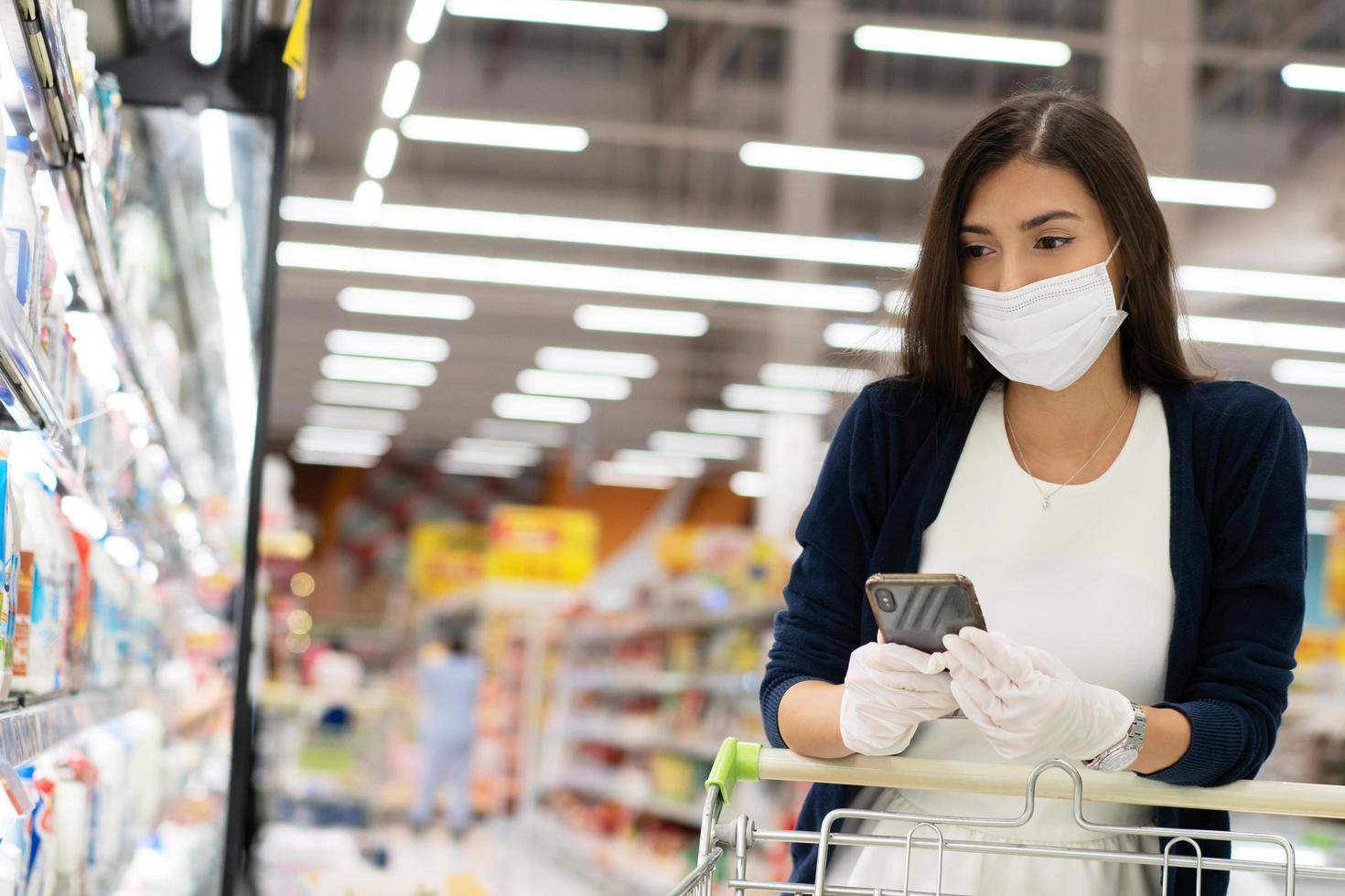 Woman wearing protective mask and rubber glove checking shopping list on mobile phone to buy stuff at grocery store. new normal lifestyle during Coronavirus pandemic photo