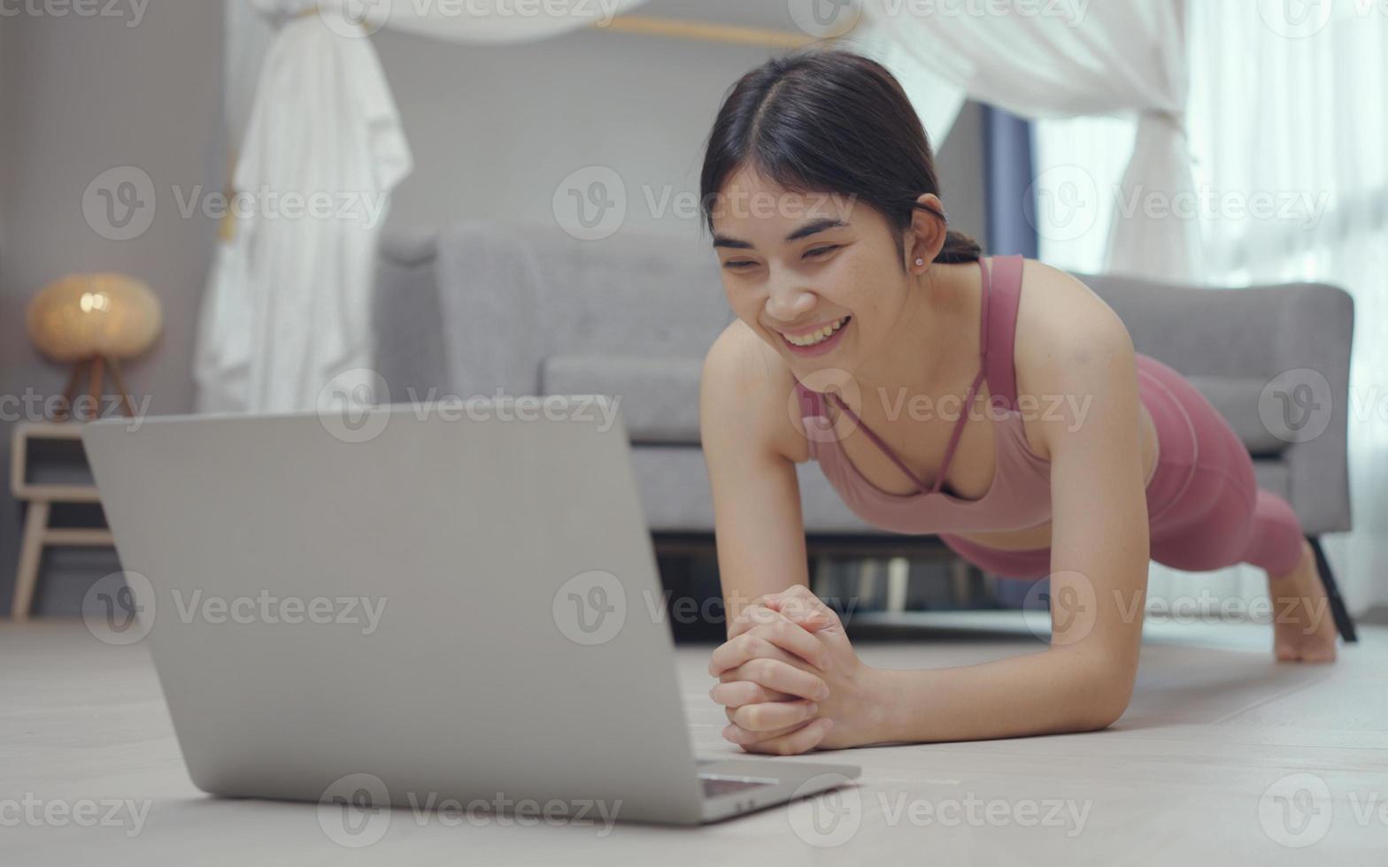 joven mujer fuerte está entrenando en casa, de pie en posición de tablón, entrenando con un entrenador en línea usando una computadora portátil. foto