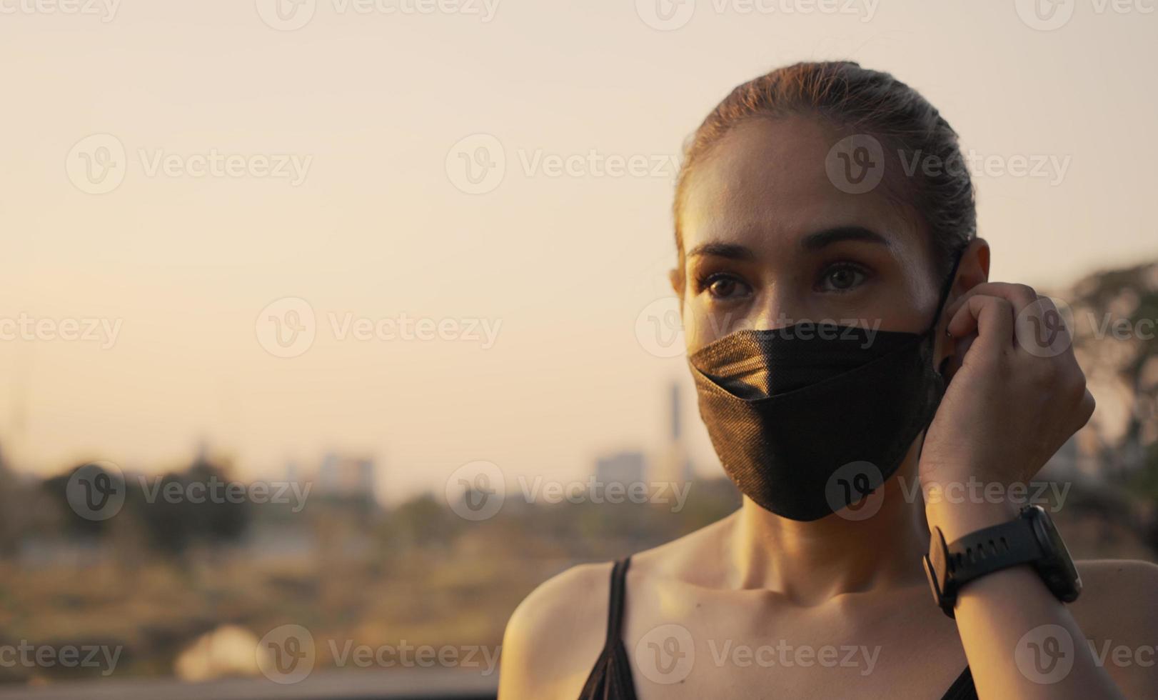 mujer con ropa deportiva usando una máscara protectora y poniendo auriculares inalámbricos antes de empezar a correr en la ciudad al atardecer. foto