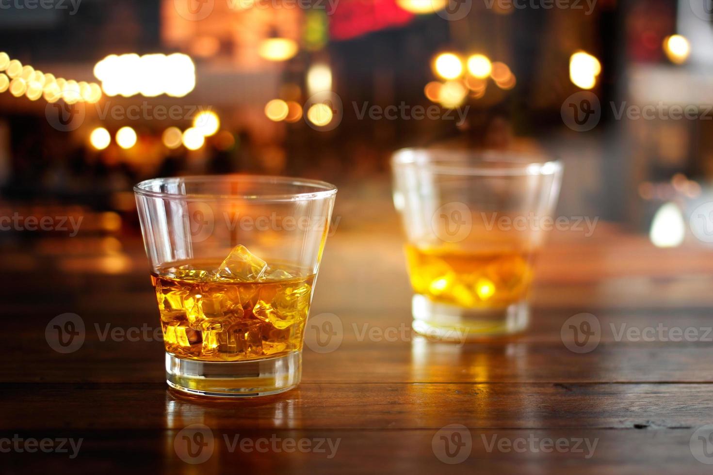 Whiskey glass drink with ice cube on wooden table in colorful night bar photo