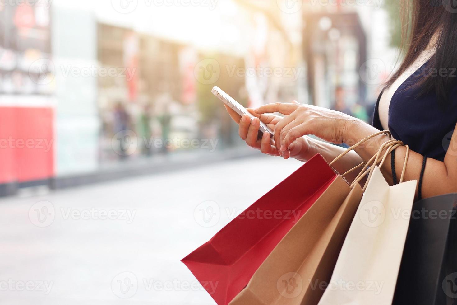 mujer usando un teléfono inteligente y sosteniendo una bolsa de compras mientras está de pie en el fondo del centro comercial foto