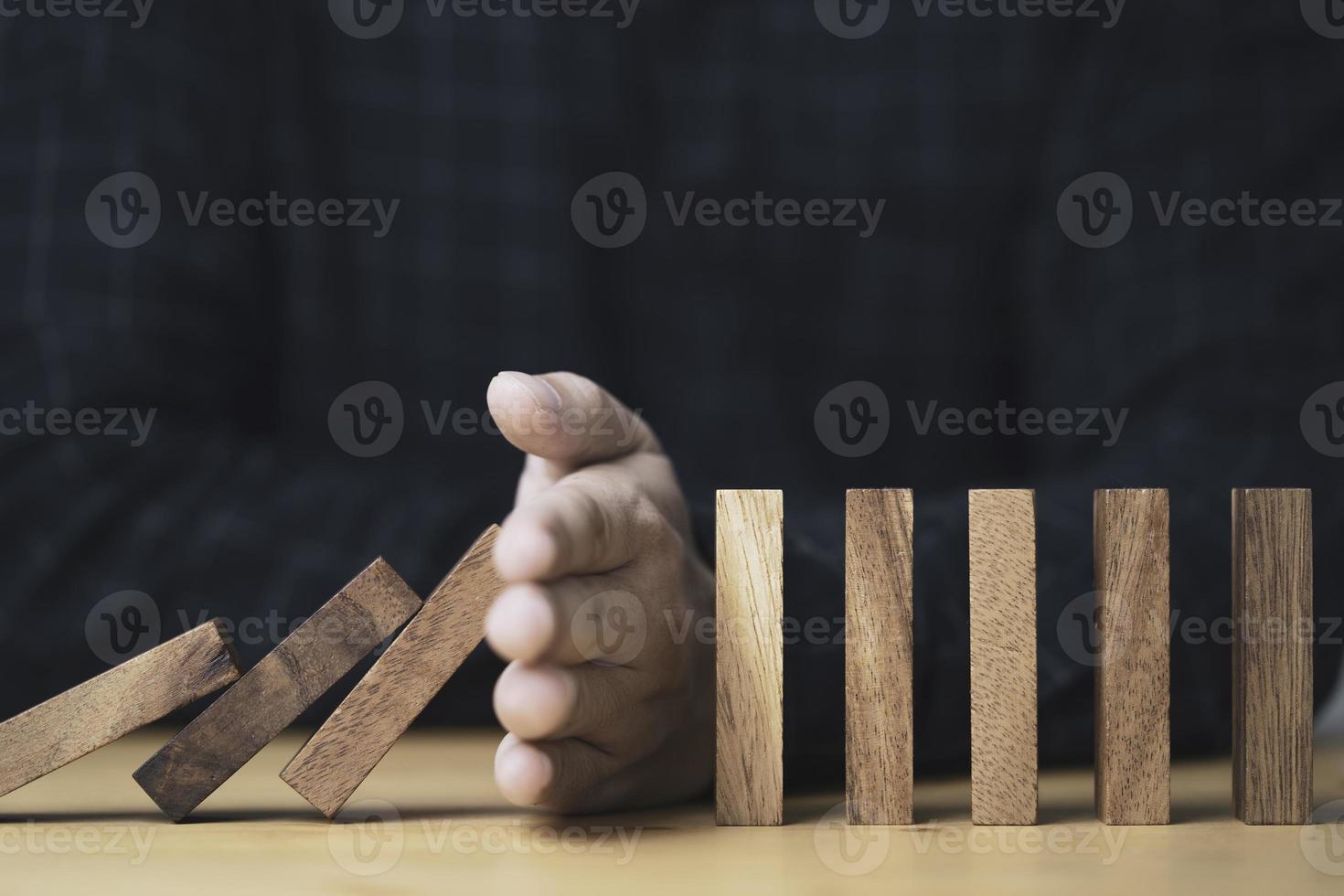 Businessman uses his hand to stop a falling wooden block to a standing wooden block domino , It is a symbol of protection against damage or stop loss for crisis management concept. photo