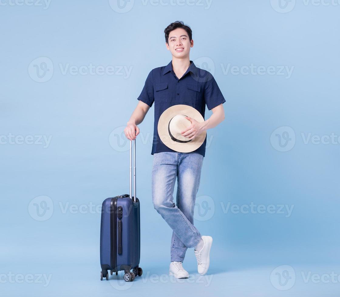 Image of young Asian man holding suitcase on blue background, travel concept photo