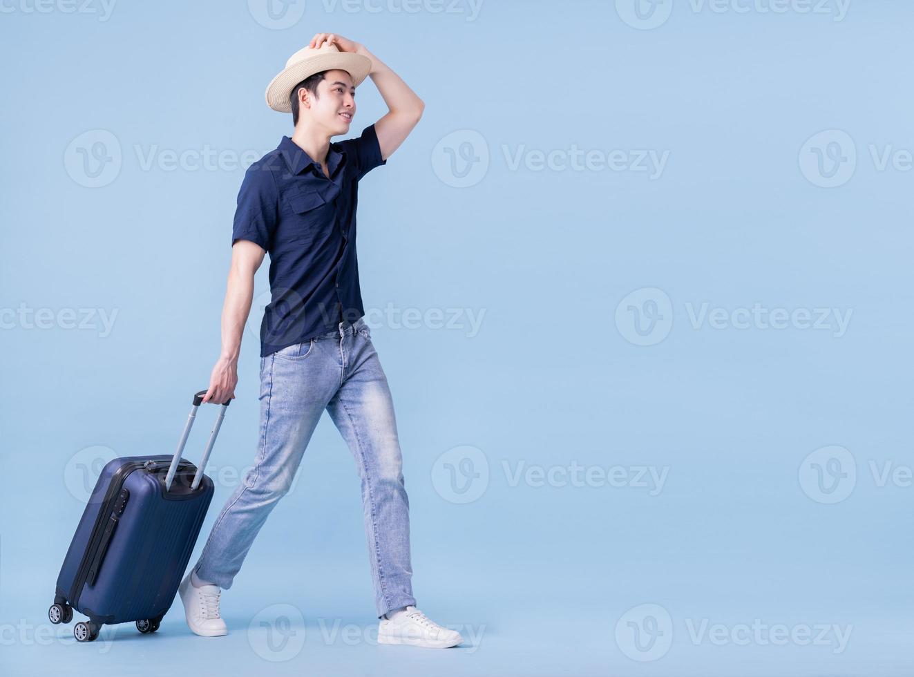 Image of young Asian man holding suitcase on blue background, travel concept photo