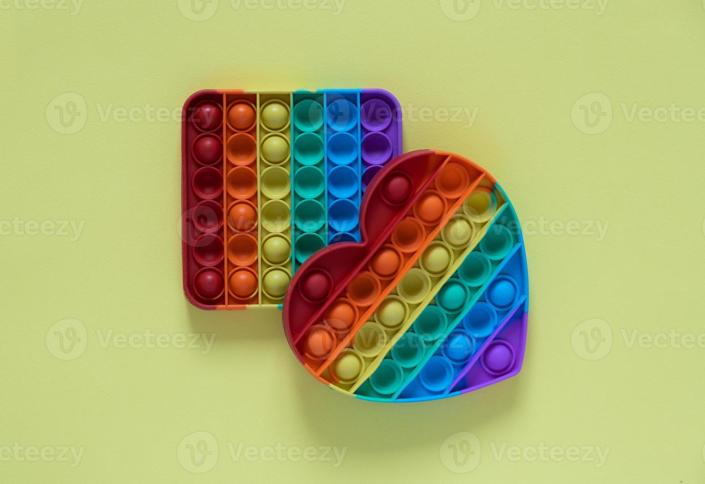 Color pop it antistress toy for children. rainbow heart shaped isolated on yellow background . 2 toys - square and in the shape of a heart. Pop it toy. photo