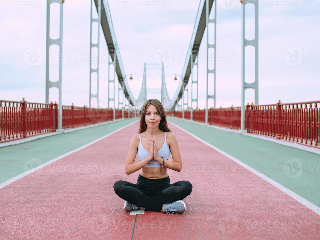 beautiful, sporty, fit, young caucasian woman in sportswear and sneakers sitting in lotus pose on the bridge. Sport, activity, yoga, urban lifestyle concept photo