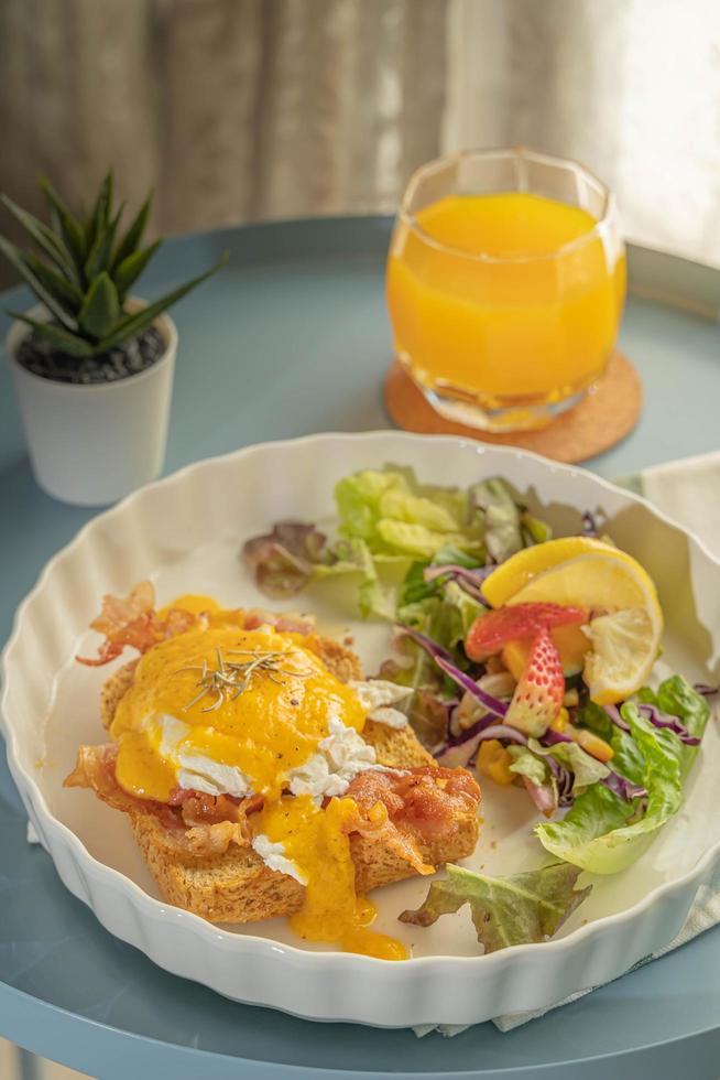 Close up on a breakfast or brunch, Eggs Benedict serve with fried bacon and toast and vegetable salad for side dish in a white plate or dish on white cloth with green strip and come with orange juice photo