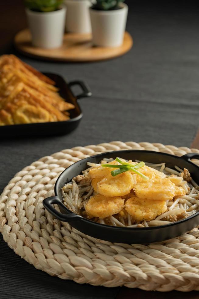 Stir-fried bean sprouts and fried egg tofu sort in black small pan on weaving  placemat, grey placemat, served with snacks, minced pork meat filled fried wontons sort in black dish. photo