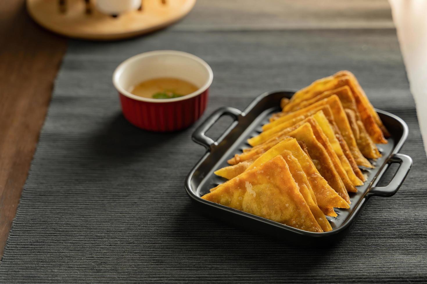 Easy recipe, snacks, minced pork meat filled fried wontons sort in black dish on grey placemat, brown wood table served with sweet plum sauce in red bowl photo