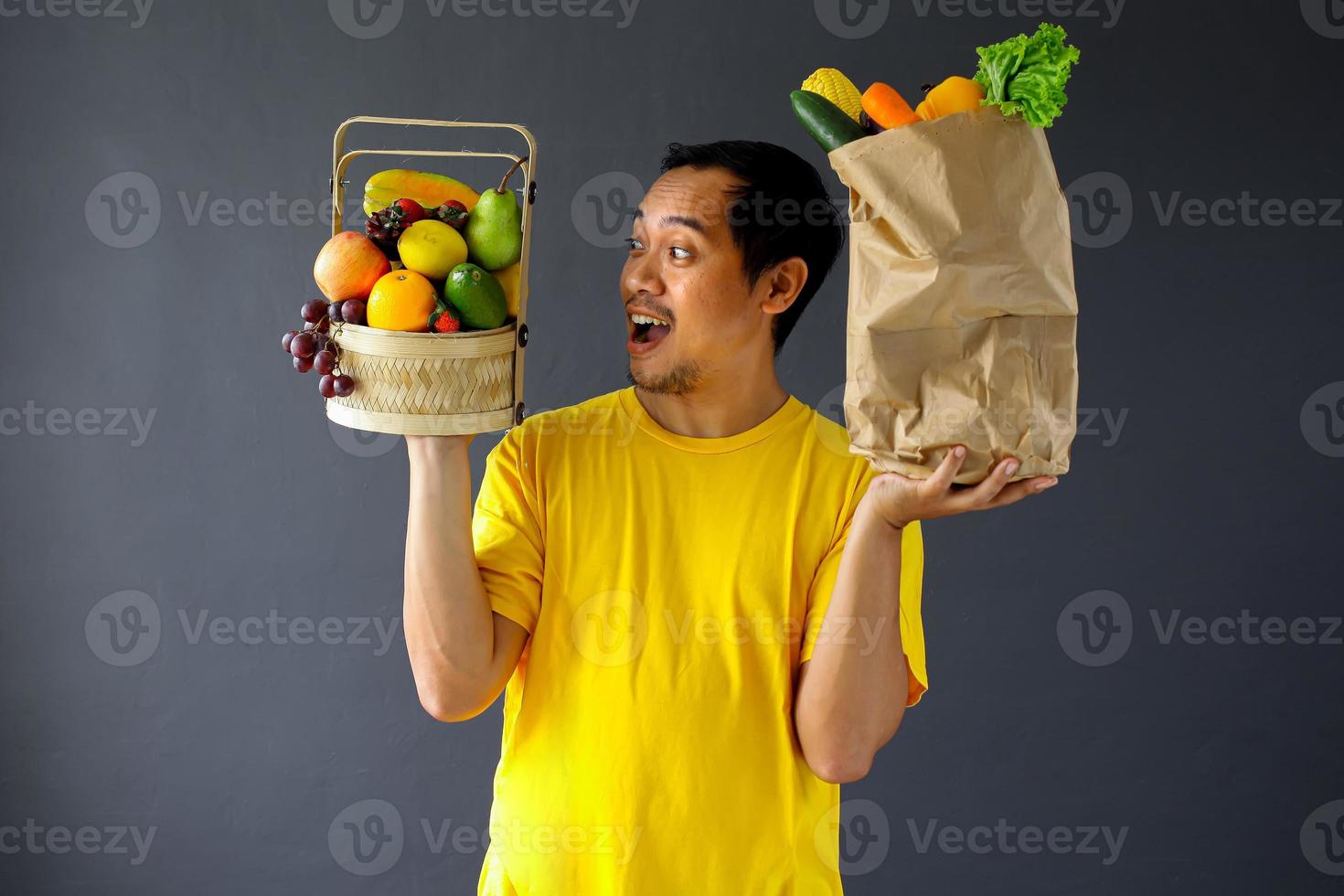 hombre asiático asombrado sosteniendo una cesta de frutas y verduras en una bolsa de compras para el concepto de campaña de estilo de vida saludable foto