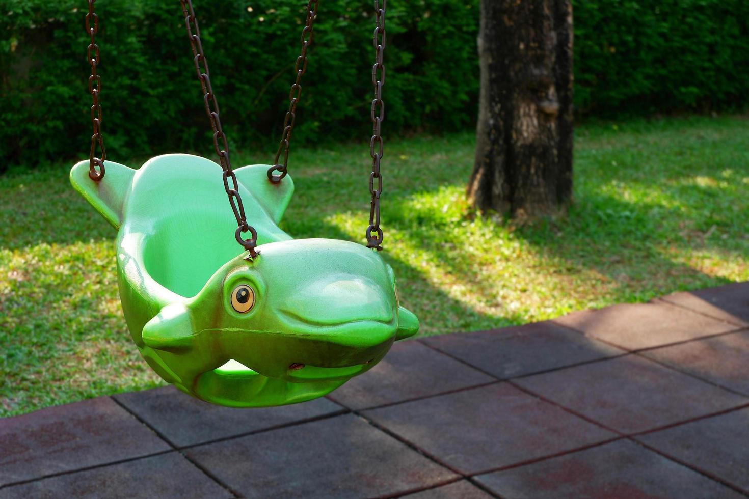 The old empty green hanging swing in dolphin shape and rubber floor tiles with green lawn in outdoor playground area at public park photo