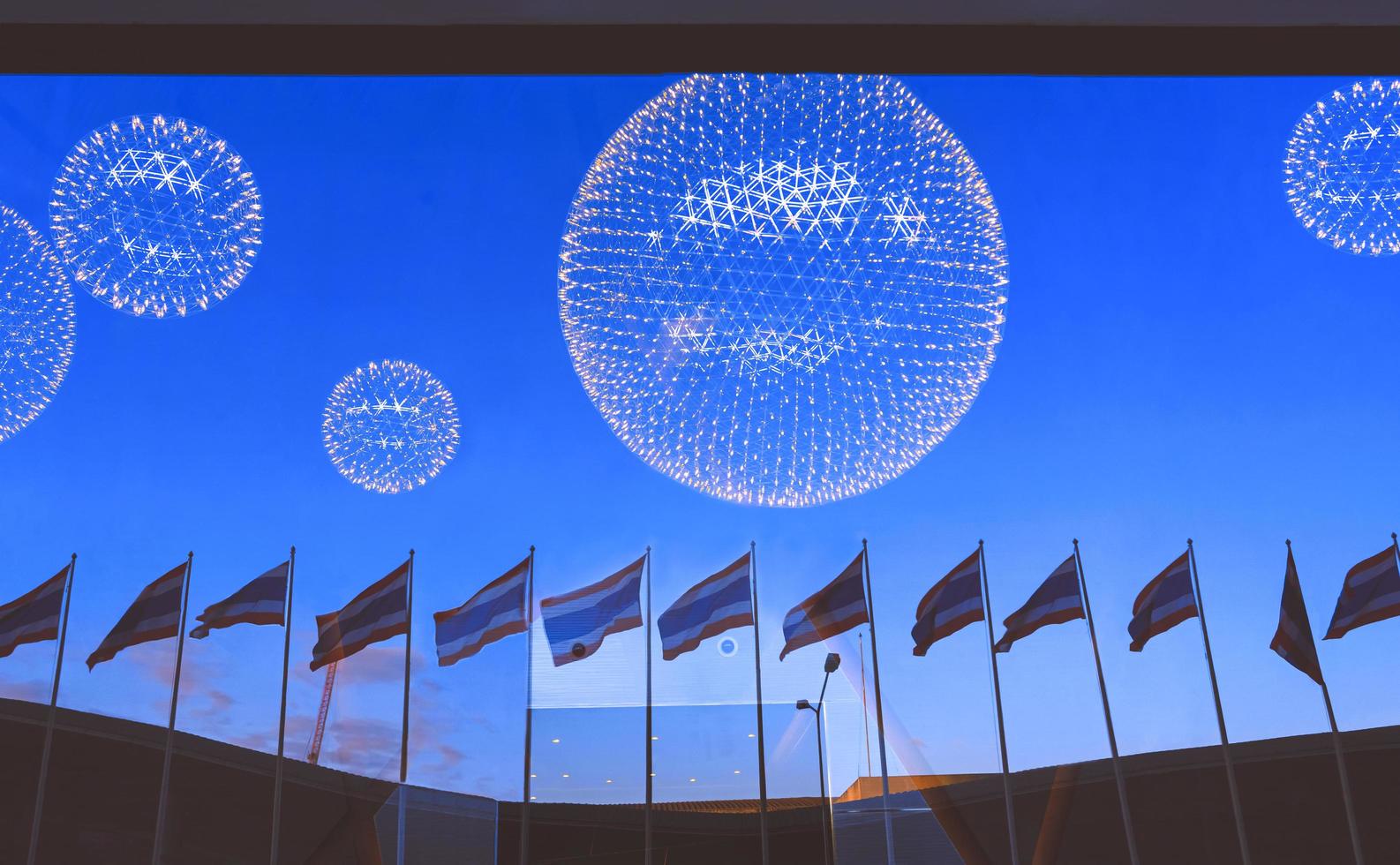 Softly focus of beautiful reflection of Thai flag poles row with curved building roof on glass wall surface and illuminated round chandeliers inside of modern building at sunset time photo