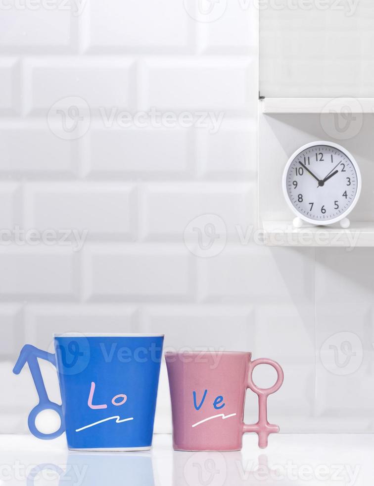 Couple blue and pink coffee mugs with round table clock on shelf in white tile wall background photo