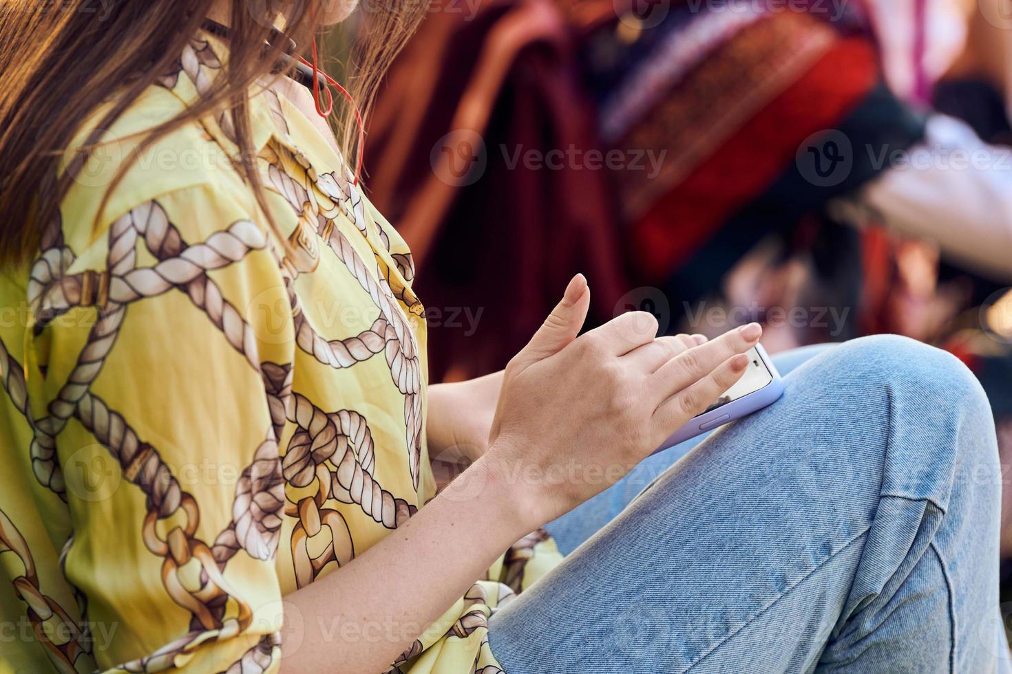 Sitting woman in yellow blouse and blue jeans using smartphone for messaging, scrolling social media photo