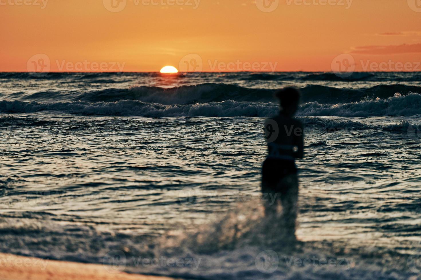 Female silhouette in blue sea waves at summer sunset, half sun below horizon, beachfront holiday photo