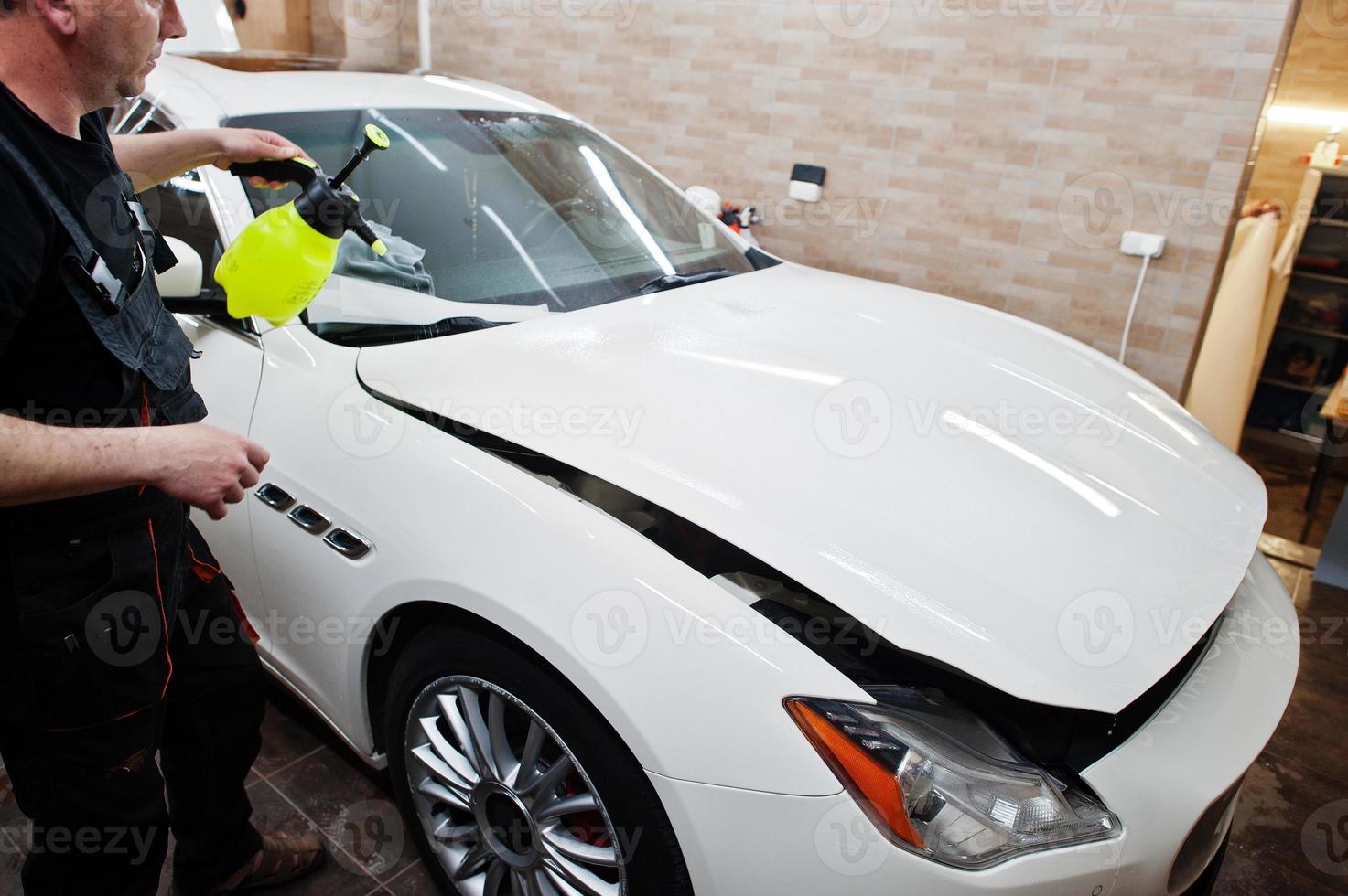 Worker in detailing garage put polyurethane anti-gravel film cover in white luxury car. photo