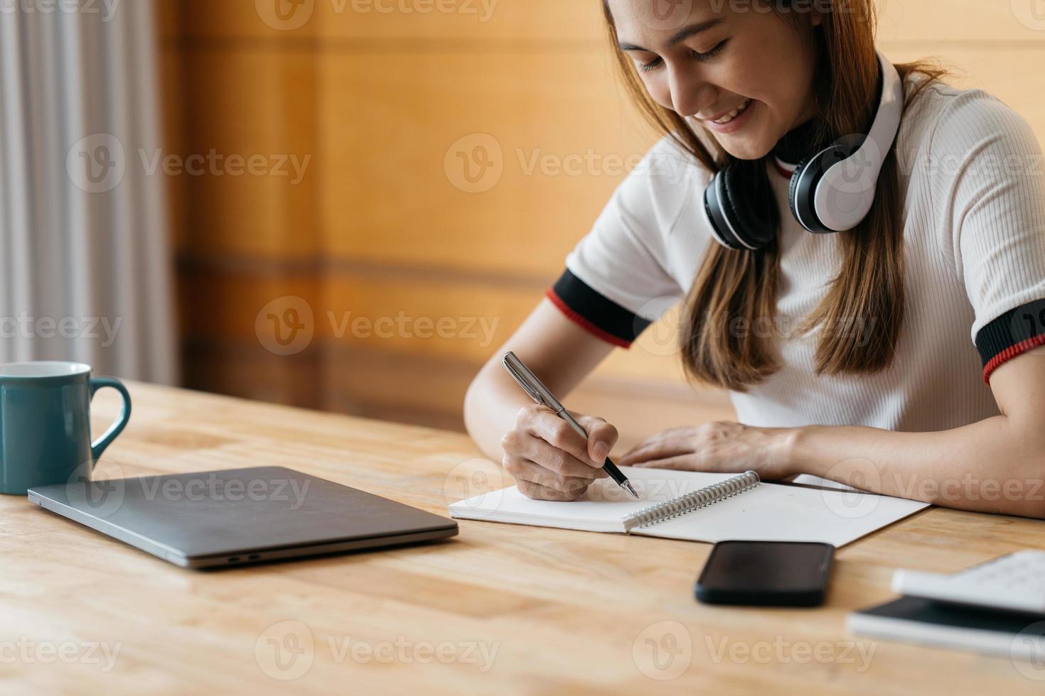 asiático feliz en el uso de auriculares portátil tomar cursos de capacitación en línea tomar notas. una joven estudiante asiática sonriente con auriculares estudia en una computadora desde casa. concepto de educación a distancia. foto