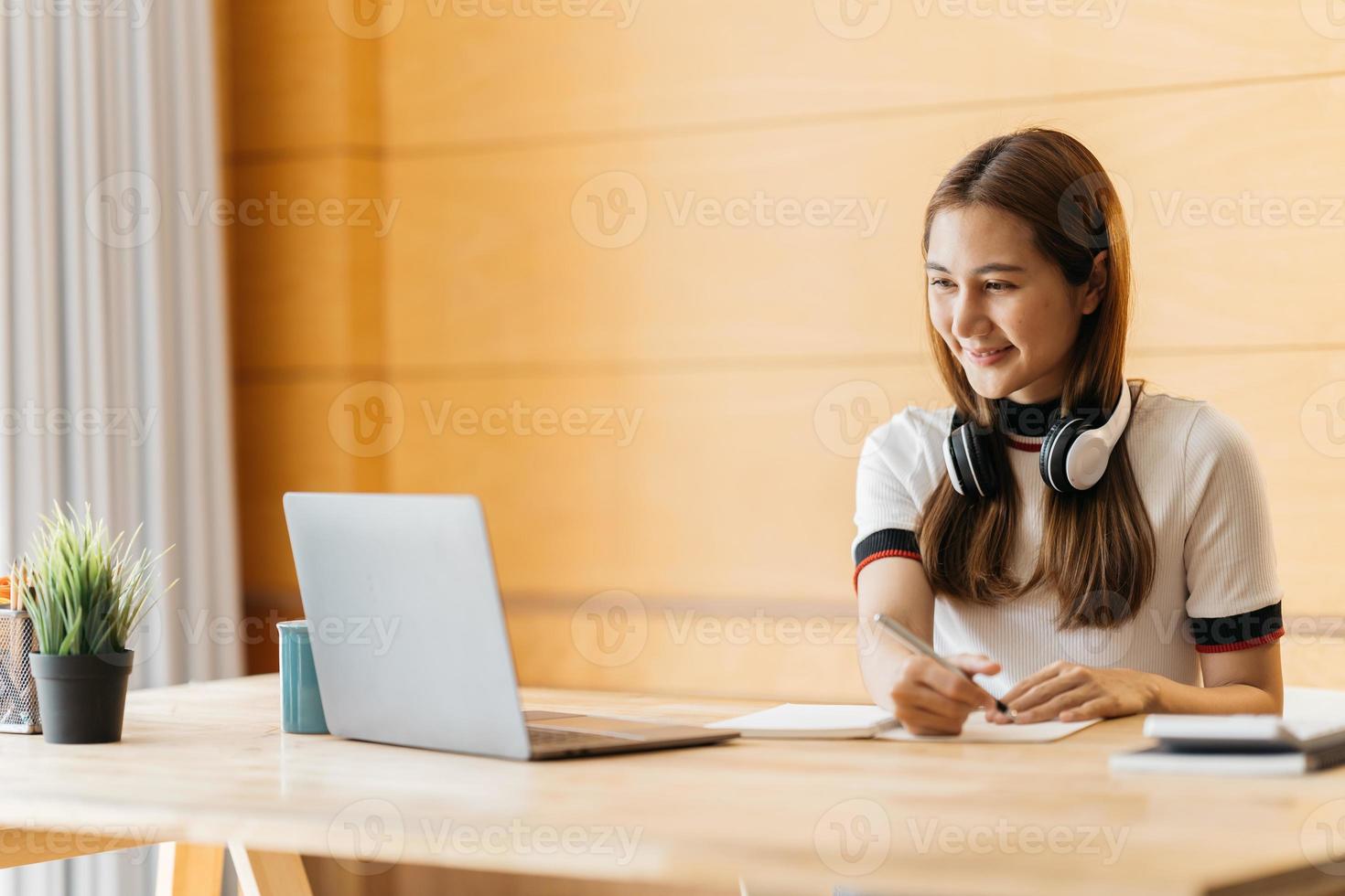 Happy asian businesswoman wearing headset make notes talk by conference call doing video chat on laptop in office, smiling chinese support service agent write information consult customer photo