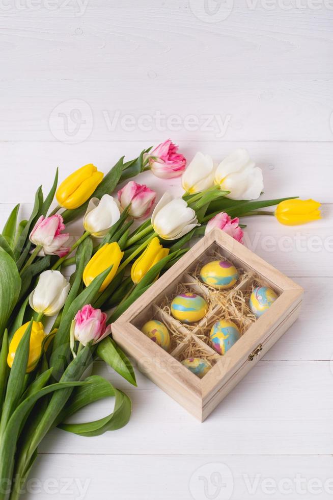 Top view of colorful easter eggs on a bed of straw in a long wooden box on a white wooden table and tulips, place for text photo