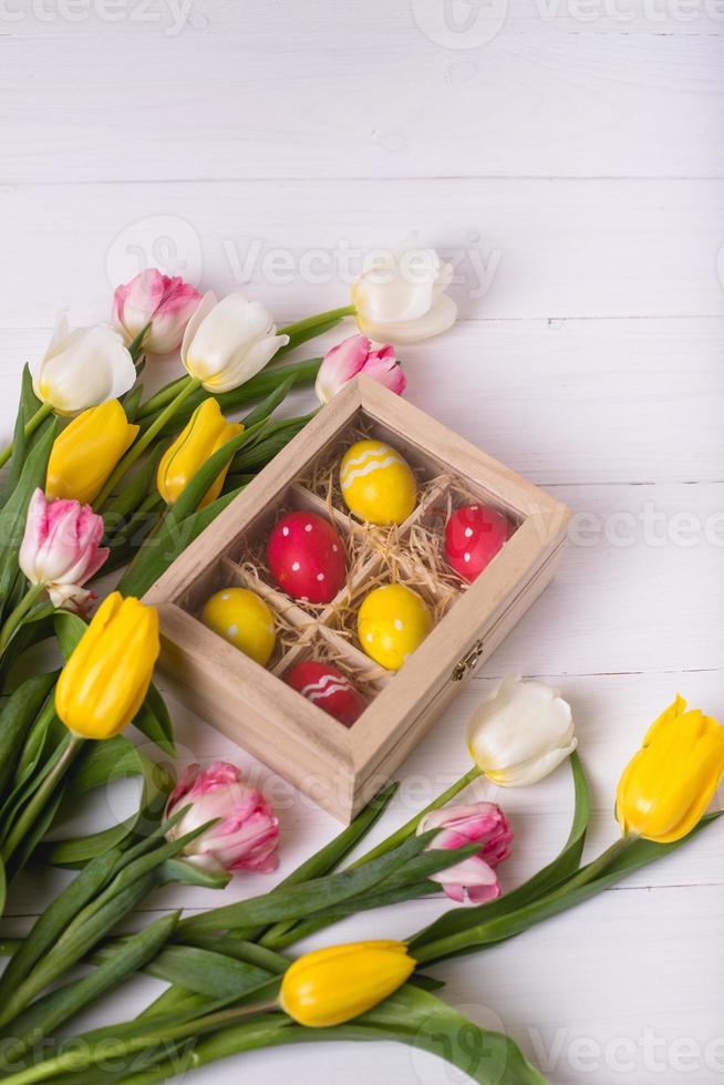 Top view of colorful easter eggs on a bed of straw in a long wooden box on a white wooden table and tulips, place for text photo