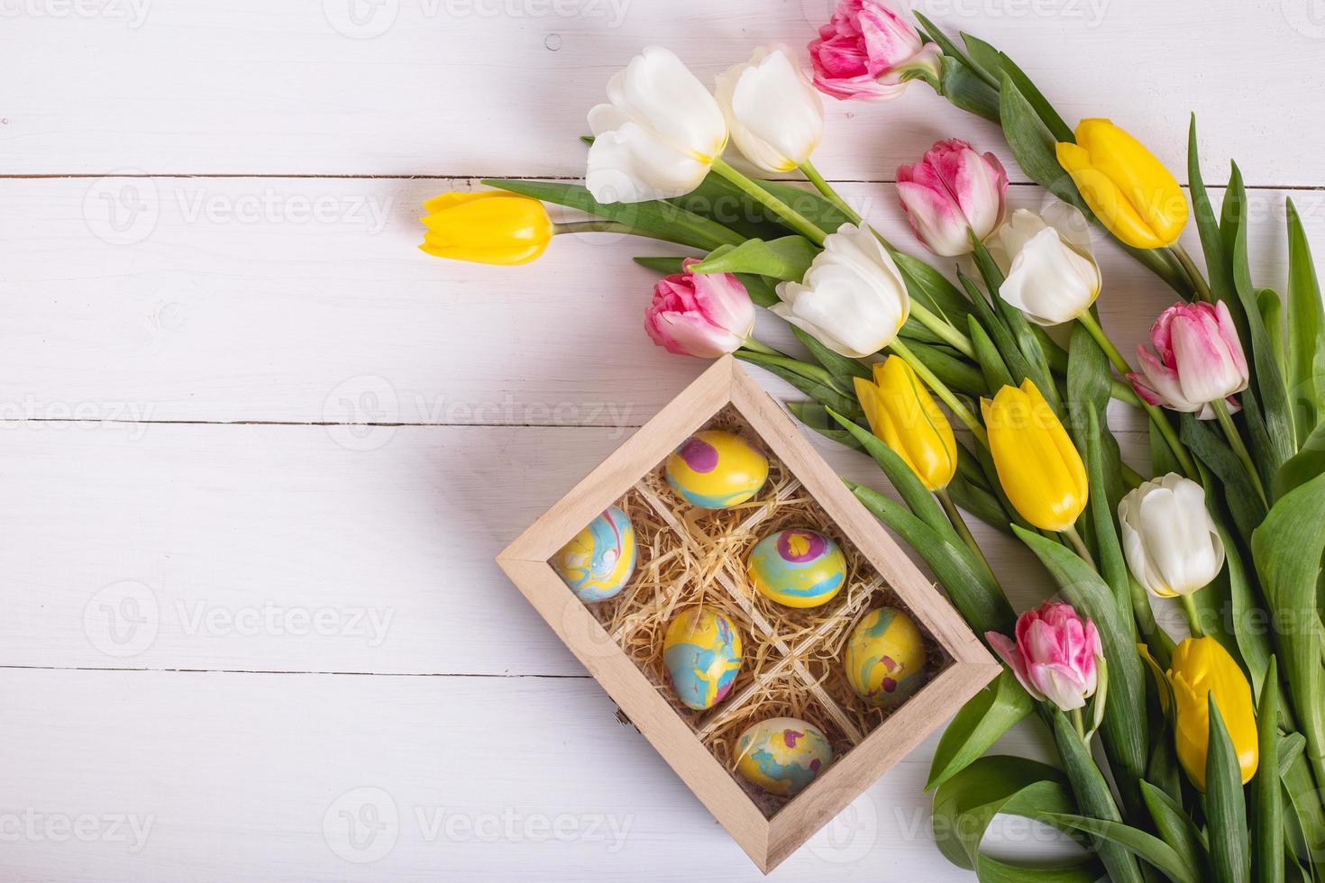 vista superior de coloridos huevos de Pascua sobre un lecho de paja en una caja de madera larga sobre una mesa de madera blanca y tulipanes, lugar para el texto foto