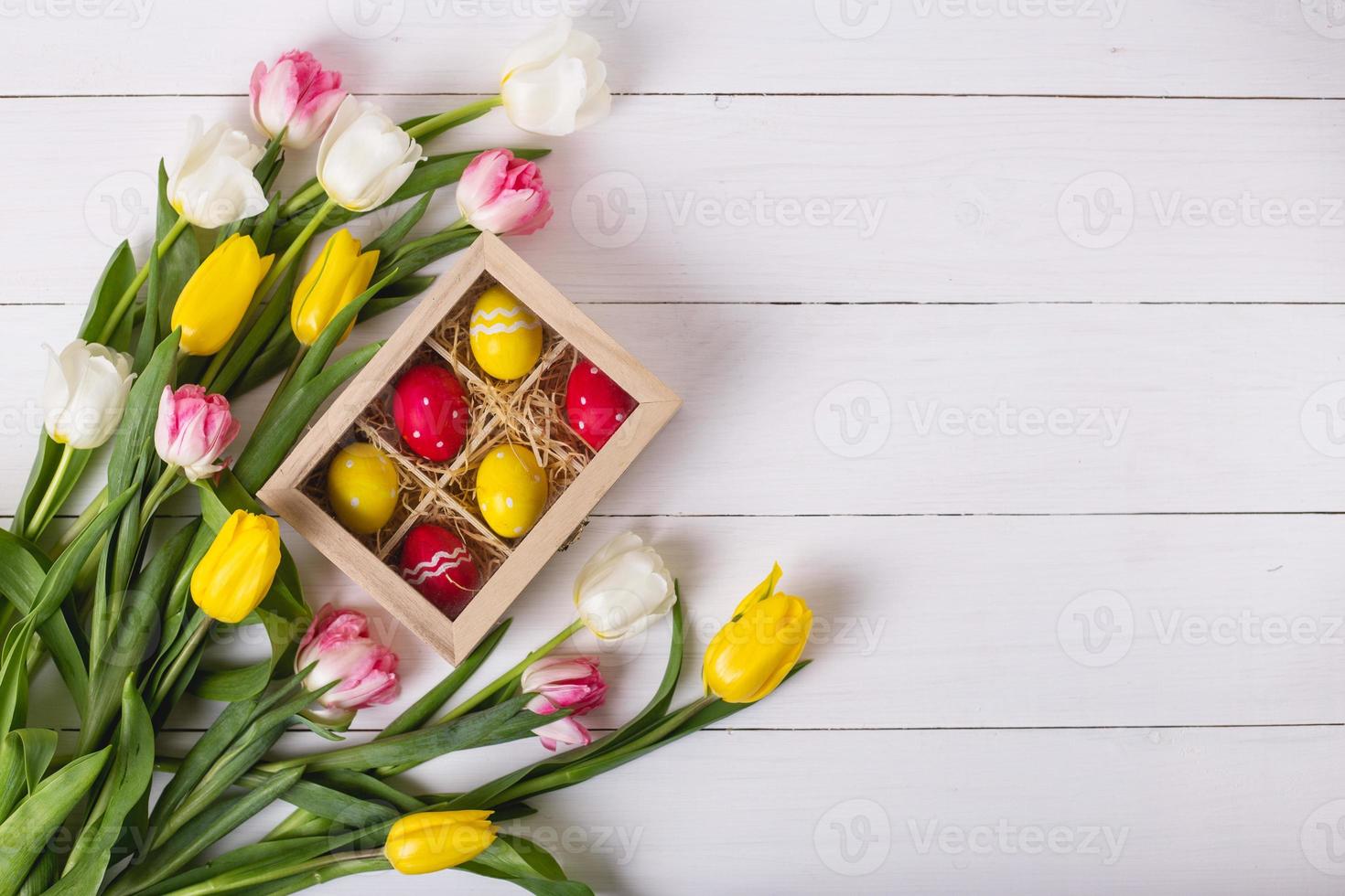 Top view of colorful easter eggs on a bed of straw in a long wooden box on a white wooden table and tulips, place for text photo