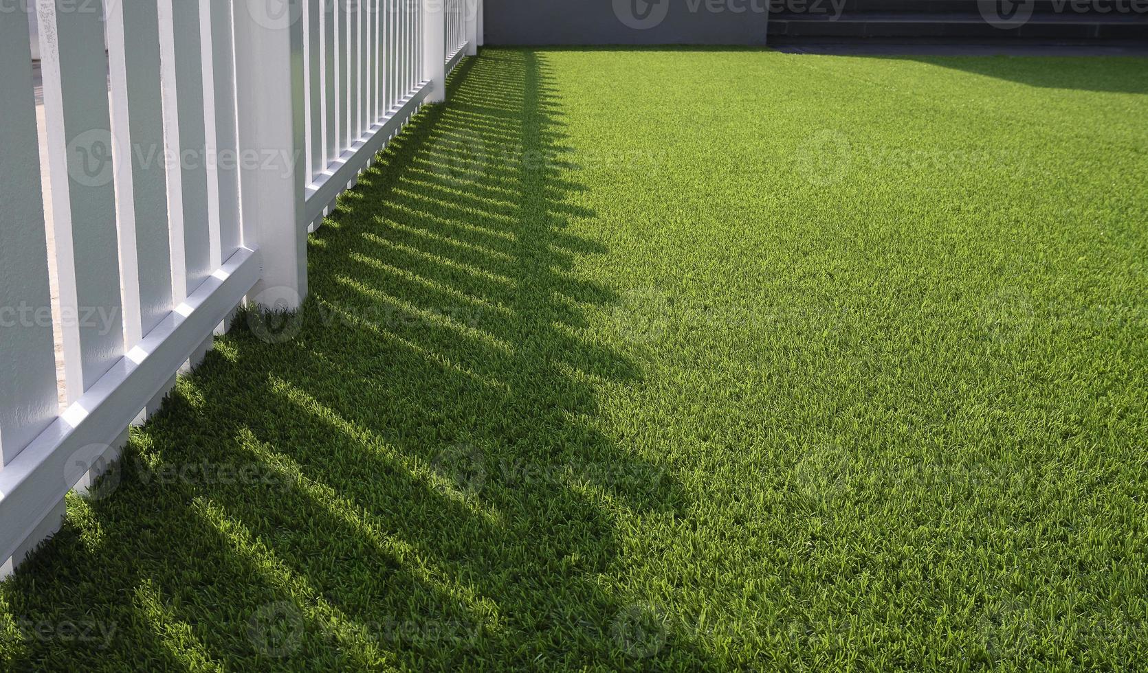 Sunlight and shadow of white wooden fence on green artificial turf surface in front yard of home, selective focus with copy space photo